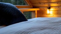 Cozy bedroom interior with soft white bedding and black pillow in wooden cabin. Warm evening light from vintage wall lamp creates relaxing atmosphere.