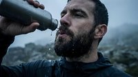 Adult male with dark beard and facial hair, appearing to be of Mediterranean or Middle Eastern descent, drinking water from a bottle outdoors in misty mountain terrain during survival or hiking activity.