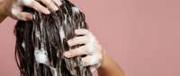 Woman applying shampoo to wet brown hair during washing routine against pink background for hair care and beauty concepts.