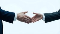 Two businessmen in dark suits extending hands for professional handshake against clean white background representing partnership and agreement.
