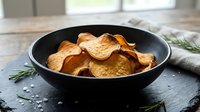 Crispy baked sweet potato chips in a dark bowl on a slate board with rosemary and sea salt, healthy snack concept.