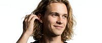 Young man with long wavy blonde hair, Caucasian ethnicity, touching his hair with hand, wearing black shirt, smiling at camera against white background for portrait use.