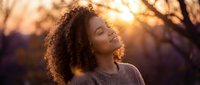 Young African American woman with curly hair enjoying peaceful moment outdoors during golden hour sunset with warm lighting and serene expression.