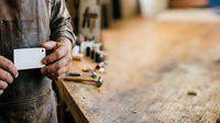 Craftsman in work apron holding blank business card at wooden workbench with tools, suitable for trade business branding and professional services marketing.