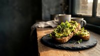 Fresh avocado toast with microgreens on sourdough bread served on slate board with coffee cup in cozy morning light for healthy breakfast concept.