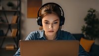 Young Caucasian woman wearing headphones working on laptop computer at home office desk, focused on screen for remote work, online learning or gaming.