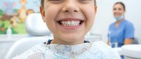 Happy young child patient smiling broadly showing healthy white teeth during dental checkup with female dentist in background at modern clinic office.