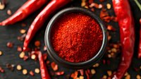 Red chili powder in black ceramic bowl surrounded by dried red hot peppers and scattered seeds on dark wooden background, top view food photography.