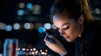 Young woman looking at smartphone at night with city lights bokeh in background, illuminating her face with blue glow.