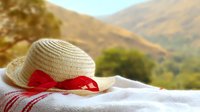 Straw sun hat with red ribbon lying on white towel against blurred mountain landscape background in warm summer afternoon light.