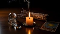 Mystical fortune telling setup with burning candle, crystal ball, tarot cards and sage bundle on wooden table creating atmospheric spiritual ambiance.