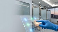 Close-up of gloved hand touching interactive digital display with holographic icons in modern office environment with glass partitions and city skyline view.