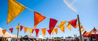 Colorful triangular bunting flags in red, orange, and yellow hang against a bright blue sky at an outdoor summer festival event.