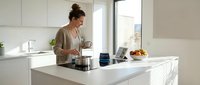 Young woman cooking in modern white kitchen with laptop on counter, preparing healthy meal at home during morning routine.