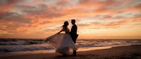 Romantic wedding couple silhouetted against dramatic sunset sky on ocean beach. Bride in flowing gown and groom embrace during golden hour ceremony.