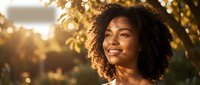 Young African American woman with curly hair smiling joyfully in warm golden sunlight outdoors during sunset, expressing happiness and positive emotions.
