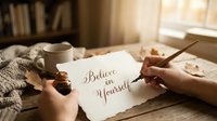 Hands writing Believe in Yourself in elegant calligraphy on textured paper, cozy autumn setting with mug and dried leaves on wooden table.