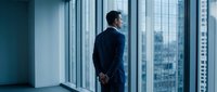 Professional businessman in dark suit standing by floor-to-ceiling windows overlooking urban cityscape, contemplating future business decisions and opportunities.
