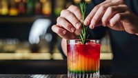 Bartender garnishing colorful layered cocktail with fresh rosemary sprig in upscale bar setting for mixology and hospitality industry.