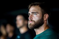 Bearded Caucasian man in green t-shirt looking thoughtfully into distance with determined expression against dark background, dramatic side lighting creates moody atmosphere.
