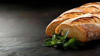 Freshly baked crusty artisan bread loaf with golden brown crust and fresh basil leaves on dark stone background, dramatic side lighting for food photography.