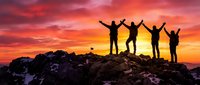 Five silhouetted figures stand on rocky summit with arms raised celebrating success against dramatic orange and red sunset sky backdrop.