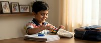 Young Hispanic boy student doing homework at desk in bright home interior with natural lighting and family photos on shelf behind.