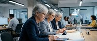 Mature women with gray hair wearing glasses sit at conference table taking notes during business meeting in modern office environment with colleagues working in background.