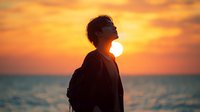 Young Asian man silhouette against dramatic orange sunset sky over ocean, looking up with peaceful expression, wearing casual clothes and backpack.