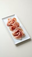 Thinly sliced cured prosciutto arranged on a white rectangular plate against a neutral background, perfect for food menus and culinary blogs.