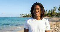 Young African American man with dreadlocks smiling on tropical beach wearing white t-shirt with palm trees and turquoise ocean in background.