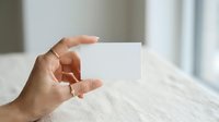 Person holding blank white card in hand against blurred interior background, suitable for business branding, marketing mockups, and promotional material presentations.