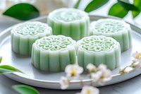 Traditional Asian snow skin mooncakes in soft mint green color with floral pattern on white ceramic plate, decorated with cherry blossoms and green leaves.
