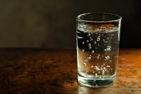 Clear glass of sparkling water with rising bubbles on rustic wooden surface against dark background, dramatic side lighting creates moody atmosphere.