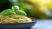 Fresh spaghetti pasta with pesto sauce and basil leaf garnish served in dark ceramic plate against blurred green background, selective focus.