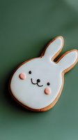 Sweet bunny-shaped sugar cookie decorated with white royal icing, pink cheeks, and simple black smile on green background. For Easter, spring celebrations.