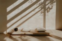 Minimalist interior with window light casting diagonal shadows on wall, white pillows and ceramic bowl on wooden shelf, creating zen atmosphere and calm mood.