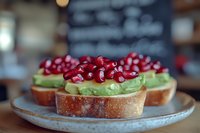 Fresh avocado toast topped with vibrant pomegranate seeds served on rustic sourdough bread, arranged on ceramic plate against blurred cafe background.
