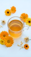 Overhead view of golden herbal tea in clear glass cup surrounded by fresh and dried yellow and orange gerbera daisies on white background, ideal for wellness and natural beverage marketing.