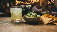 Refreshing cocktail with lime and mint served alongside guacamole dip and tortilla chips on wooden table with warm bokeh lights in background for bar, restaurant, and beverage marketing.