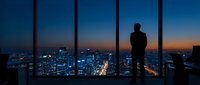 Businessman silhouette standing at office window overlooking illuminated city skyline at dusk. Corporate success and urban planning concept for presentations.