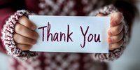 Female hands in cozy knitted sweater sleeves holding white paper note with handwritten Thank You message against blurred background, warm winter gratitude concept.