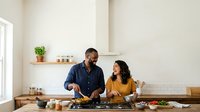 Man and woman cooking together in modern kitchen, preparing fresh ingredients on wooden counter with stainless steel cookware and utensils for meal preparation and culinary instruction.