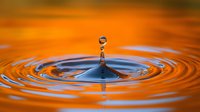Single water drop creating concentric ripples against vibrant orange sunset reflection, captured in high-speed macro photography showing surface tension and liquid dynamics.