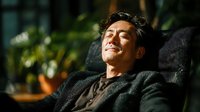 Relaxed middle-aged Asian man with closed eyes enjoying peaceful moment sitting in comfortable chair with warm sunlight streaming across face.