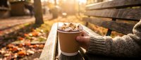 Hands holding warm hot chocolate with marshmallows on wooden bench during golden autumn sunset in park with fallen leaves creating cozy seasonal atmosphere.