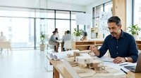 Architect or designer with dark hair and beard, wearing navy blue shirt, working on architectural models and blueprints at wooden desk in modern office with large windows and colleagues in background.