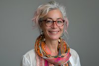 Confident mature Caucasian woman with silver hair, glasses and colorful scarf smiling warmly against grey background. Professional portrait for business or lifestyle.