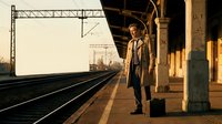 Young businessman in coat waiting on vintage train station platform with suitcase during golden hour sunset. Urban commuter travel and transportation concept.