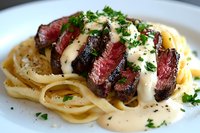 Sliced medium-rare steak served over fettuccine pasta with creamy white sauce and fresh chopped parsley garnish on white plate. Gourmet restaurant presentation.